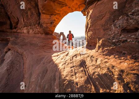 Una donna che si trova in un passaggio attraverso una parete di arenaria dove, dall'altro lato, si vede il delicato Arco all'Arches National Park, Utah, USA. Foto Stock