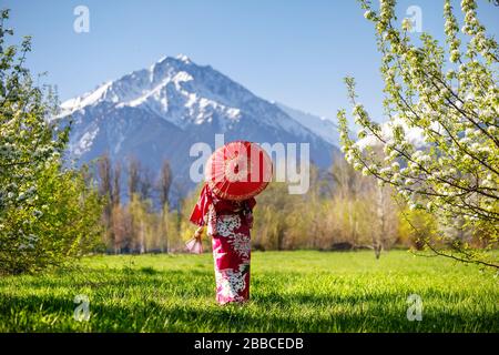 Donna in kimono con ombrellone rosso nel giardino con fiori di ciliegio a sfondo di montagna Foto Stock