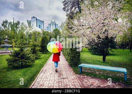 Woman in Red coat con ombrello arcobaleno passeggiate nel parco con alberi in fiore al cielo nuvoloso Foto Stock