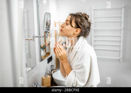 Giovane donna in accappatoio che si prende cura della sua pelle, guardando lo specchio nel bagno. Cura della pelle del viso e concetto di benessere Foto Stock