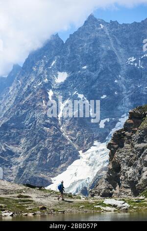 Breuil-Cervinia, Italia - 23 luglio 2019: Viaggiatore con zaino trekking da solo in montagna. Backpacker passeggiata collina percorso vicino lago di montagna con collina rocciosa sullo sfondo. Concetto di alpinismo. Foto Stock