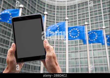 Mani di un uomo irriconoscibile che tiene un tablet a schermo vuoto all'aperto davanti a una fila di bandiere dell'Unione europea Foto Stock