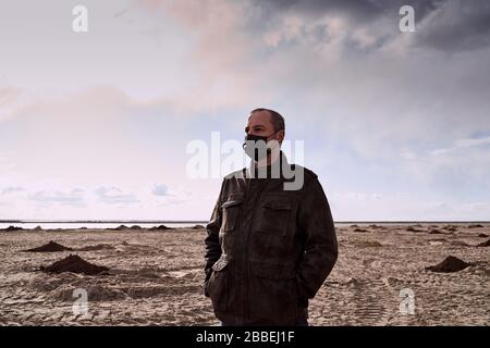 Apocalisse e cataclisma dovuti al coronavirus. Uomo maturo in maschera nera e cappotto nero si trova in mezzo al deserto, Sands. Solo, perso. Paesaggista surreale Foto Stock