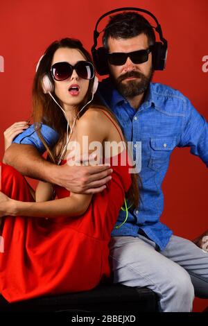 Relax, festa e musica concetto. Coppia in amore indossa cuffie e occhiali da sole. Uomo con barba e ragazza abbracciano su sfondo rosso. Gli appassionati di musica con volti tranquilli amano la musica. Foto Stock