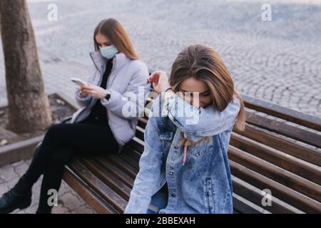 Ragazza in maschere. Tema del coronavirus. Le donne in città si siedono sulla panchina senza maschera protettiva, durante la quarantena. Le donne starnutiscono e tossiscono Foto Stock