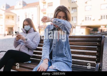 Ragazza in maschere. Tema del coronavirus. Le donne in città si siedono sulla panchina senza maschera protettiva, durante la quarantena. Le donne starnutiscono e tossiscono Foto Stock