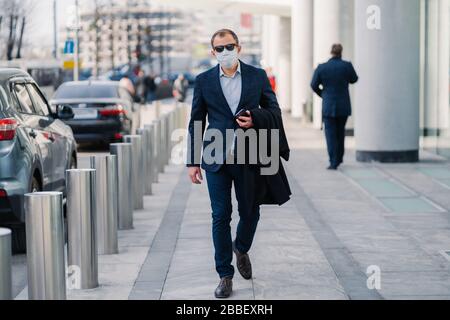 Un uomo d'affari indossa una maschera medica protettiva, cammina nella strada della città, vestita con abiti formali, tiene il telefono cellulare, andando a lavorare. Salute Foto Stock