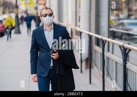 Colpo esterno di uomo d'affari in abiti formali, tiene il telefono cellulare moderno in mani, cammina per strada, si protegge con maschera medica, impedisce trasmi Foto Stock