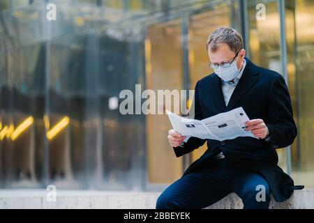Un colpo all'aperto di un capo uomo serio si rompe dopo aver camminato, legge un giornale, indossa occhiali per una buona visione, maschera medica per proteggersi da vi Foto Stock