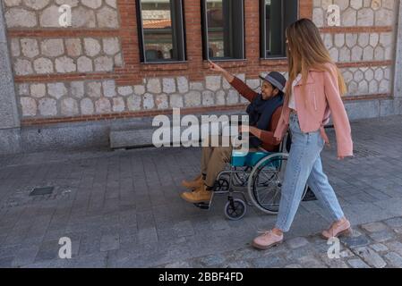 Giovane uomo latino-americano in sedia a rotelle accanto a due giovani ragazze caucasiche felici e sorridenti mentre camminano per la strada Foto Stock