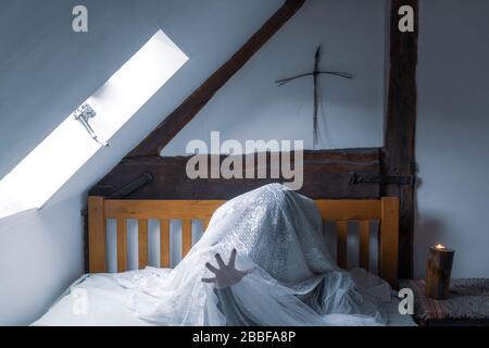 Una figura spettrale e spettrale seduta su un letto, con una mano che raggiunge con una croce sopra il letto. Foto Stock