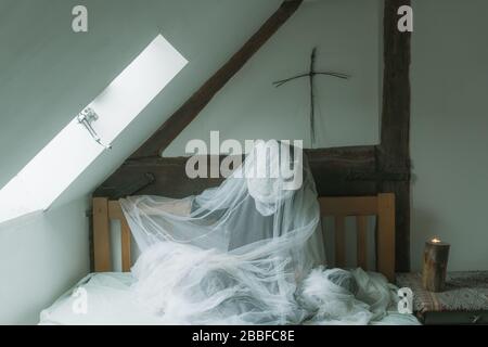 Una figura spettrale e spettrale seduta su un letto, coperta in un foglio con una croce sopra il letto. Foto Stock