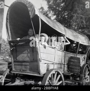 Anni '50, storico, un padre con i suoi tre figli seduti in un vecchio carro coperto di legno, del tipo usato dalla gente durante i giorni di viaggio i sentieri del selvaggio West americano. In epoca pre-industriale, il carro coperto era la forma dominante di trasporto utilizzato dalle persone che si spostano o migrano in questa epoca. Con la sua robusta struttura in legno e le robuste ruote, fu in grado di affrontare le strade e i sentieri accidentati e non sviluppati degli stati orientali. È un simbolo iconico e romantico dell'Occidente americano. Foto Stock