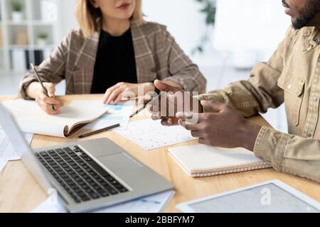 Primo piano di colleghi multietnici seduti al tavolo con documenti e dispositivi e discussione del progetto di avvio Foto Stock