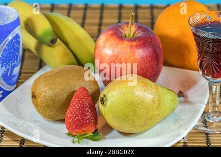 Frutta fresca tropicale e subtropicale in un grande piatto bianco. Bicchiere Cristal di vino rosso e calice di cristallo intagliato blu e bianco su piedistallo di bambù in vimini Foto Stock