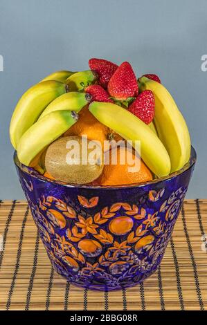 Frutta fresca tropicale e subtropicale in un grande vaso di cristallo blu. Stand di bamboo in vimini sul tavolo da pranzo, sfondo grigio. Foto Stock