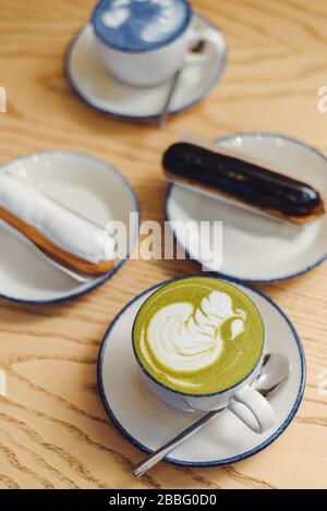 Matcha blu e verde in bicchieri su un tavolo di legno in una caffetteria accanto ad un dolce dessert. Eclairs con ganache di cioccolato e glassa con diversi condimenti. Foto Stock