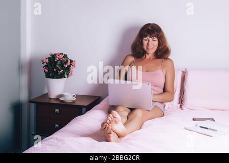 Felice donna di mezza età che lavora a casa su un computer portatile seduto sul letto in casa. Lavoro da casa concetto. Autoisolamento. Foto Stock