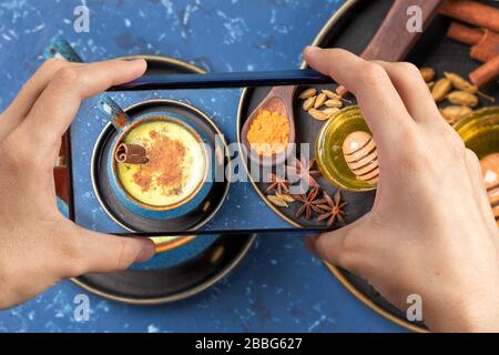 Smartphone femminile con foto sopra la tazza blu di tradizionale ayurvedico indiano sano bere latte curcuma dorato con ingredienti Foto Stock