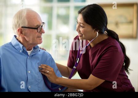 Sorridente uomo anziano che ha una conversazione con un'infermiera femminile mentre ascolta il suo battito cardiaco attraverso uno stetoscopio. Foto Stock