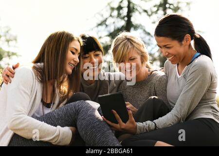 Quattro amiche felici che guardano un tablet digitale insieme in un parco soleggiato Foto Stock