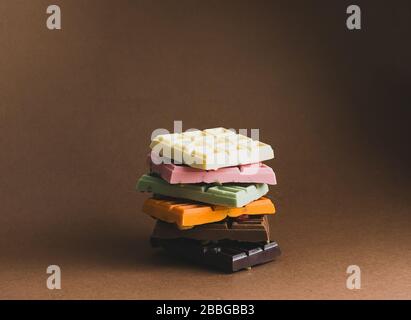 Cioccolato di vari colori e ripieni colorati con pezzi di frutta secca è accatastato su uno sfondo marrone. Vista frontale Foto Stock