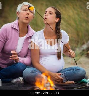La donna matura e la sua figlia incinta soffiano su un marshmallow bruciante mentre hanno un falò alla spiaggia Foto Stock