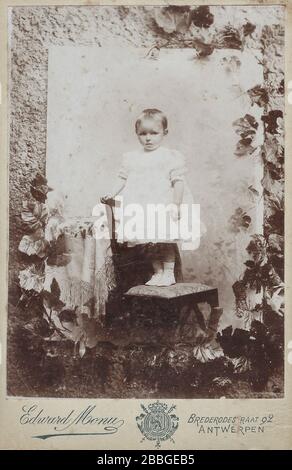 cabinet card da circa 1905 di ragazza molto giovane in abito bianco in piedi su una sedia in uno studio di fotografia; foto di una foto decorata da piante e Foto Stock