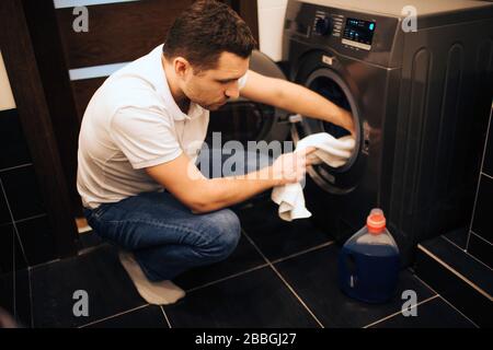 Immagine di uomo serio sicuro che mette alcuni vestiti nella lavatrice. Farcela da sola nella vasca da bagno. Mettere gli abiti all'interno. Guarda il flacone con Foto Stock