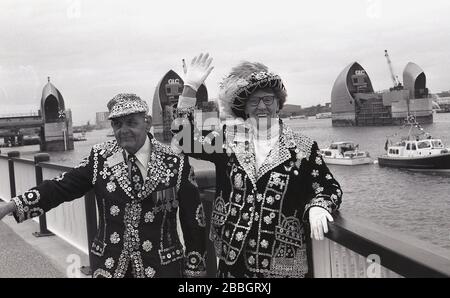 1984, il Pearly King e la Queen di Londra indossano i loro abiti tradizionali. Giacche decorate con bottoni di madreperla, in piedi insieme ai Royal Docks, all'apertura della Regina Elisabetta II della nuova barriera del Tamigi, Londra meridionale, Inghilterra, Regno Unito. Che rappresenta la cultura della classe operaia londinese, "Pearlies" come sono anche conosciuti, Si è evoluto dai Re Coster e dalle Regine, che sono stati eletti come leader dei commercianti di strada di Londra. Foto Stock