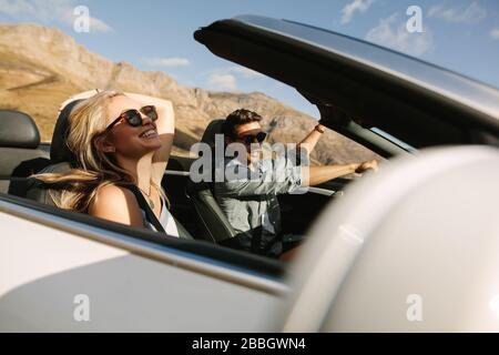 Coppia allegra che si diverte durante un viaggio in auto. Giovane uomo che guida un'auto convertibile con donna sorridente sul sedile del passeggero. Foto Stock