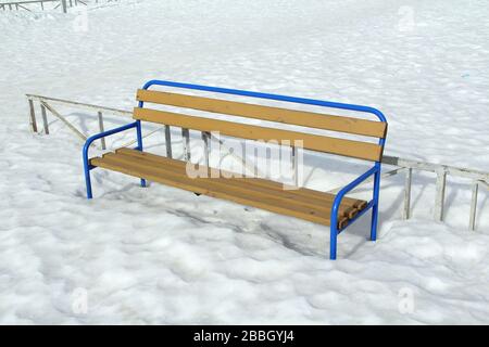 Panca leggera in legno con base blu in metallo su fondo neve. Paesaggio cittadino, area ricreativa. Stock foto per web e stampa. Foto Stock