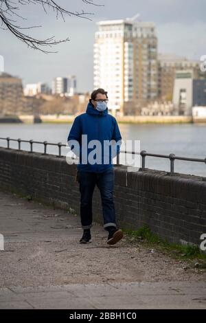 A metà degli anni '40 uomini indossano giacca blu e maschera facciale passeggiate lungo il Tamigi, con il quartiere finanziario di Londons e Canary Wharf sullo sfondo. Foto Stock