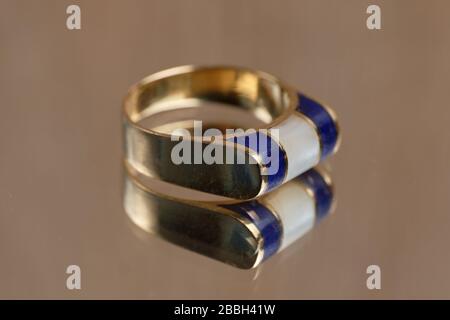 Anello dorato creativo con pietra bianca e blu su un tavolo di vetro. Primo piano. Foto Stock