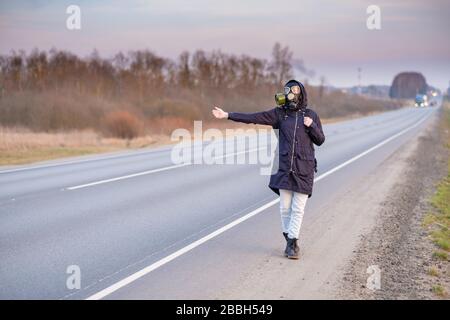 Una ragazza in abiti scuri, una maschera a gas e una cappa sulla testa. Giorno nuvoloso nel paese. Alza la mano e cerca di fermare la vettura che passa. Fuga Foto Stock