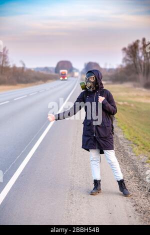 Una ragazza in abiti scuri, una maschera a gas e una cappa sulla testa. Giorno nuvoloso nel paese. Alza la mano e cerca di fermare la vettura che passa. Fuga Foto Stock