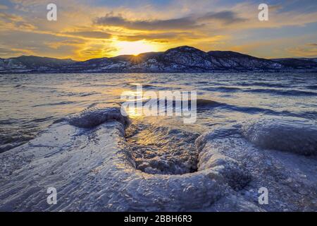 Sole che si trova dietro le montagne sul Lago Okanagan in inverno. Penticton, British Columbia, Canada. Foto Stock