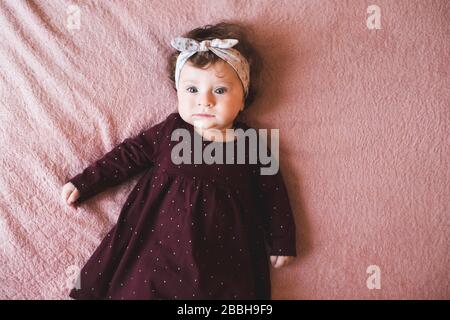 Elegante bambina di meno di 1 anno che indossa un abito alla moda e un fasciatesta sdraiato sul letto con copertura rosa. Vista dall'alto. Guardando la fotocamera. Infanzia. Foto Stock