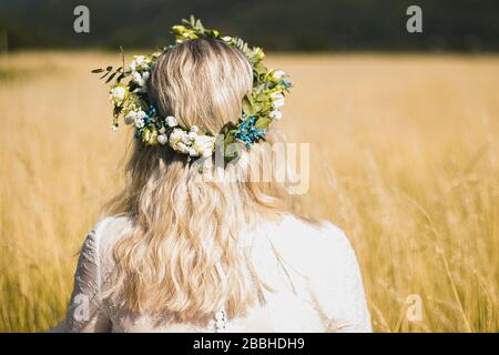 Retro della giovane donna in un abito bianco in stile boho con una corona floreale in estate nel campo. Foto Stock