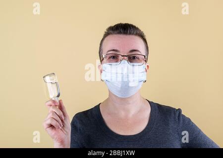 Donna adulta caucasica dai capelli molto corti nel maglione e con occhiali e maschera medica sul suo viso tiene in mano un vasetto di gel antisettico. Foto Stock