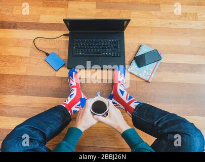 Uomo che indossa una calza jack a bandiera a Unione seduta su pavimento in legno con laptop. Lavorare da casa, auto isolamento, allontanamento sociale, Coronavirus... concetto Foto Stock