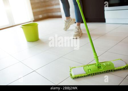 Giovane donna in cucina durante la quarantena. Pavimento di pulizia con mop verde. Vista con taglio basso. Rendere il pavimento più pulito. Secchio verde con acqua dietro. Foto Stock