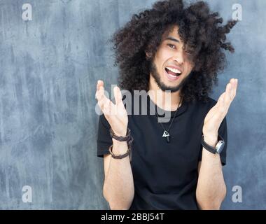 Un giovane uomo con un'acconciatura afro e una barba sta ridendo e battendo su uno sfondo grigio. Stile di strada. La gioia dei giovani. Foto Stock