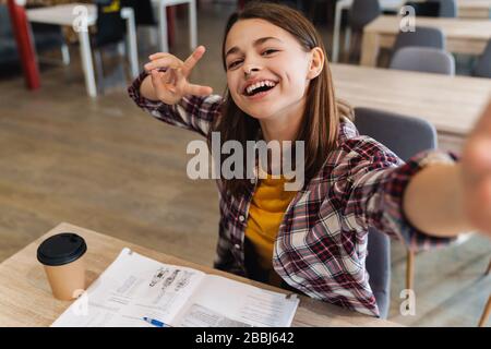 Immagine di ragazza gioiosa che prende selfie e gesturing segno di pace mentre fa i compiti nella biblioteca dell'università Foto Stock