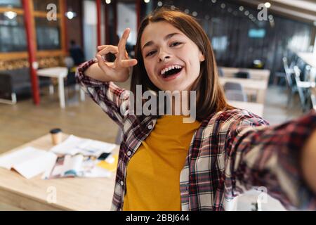 Immagine di ragazza gioiosa che prende selfie e gesturing segno di pace mentre fa i compiti nella biblioteca dell'università Foto Stock