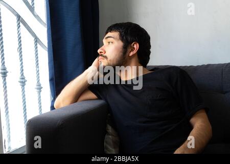 Un giovane ispanico seduto su un divano molto pensieroso che guarda fuori una finestra Foto Stock