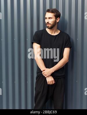 Un uomo ippopotere barbuto in una T-shirt nera e pantaloni che posano contro una recinzione metallica in un giorno estivo. Foto Stock