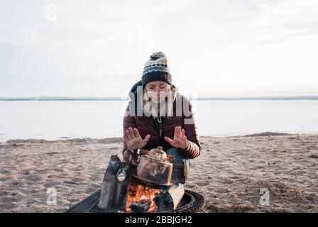 donna che scalda le mani su un fuoco di campo in attesa che l'acqua bollire Foto Stock