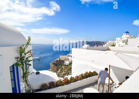 Due turisti in una terrazza laterale con vista sul mare e sulla caldera con la cupola blu in vista a Santorini, Grecia. Foto Stock