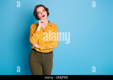 Foto di simpatica simpatica ragazza braccio dito sul mento sguardo vuoto spazio candida persona creativa indossare camicia gialla pantaloni verde isolato blu brillante Foto Stock